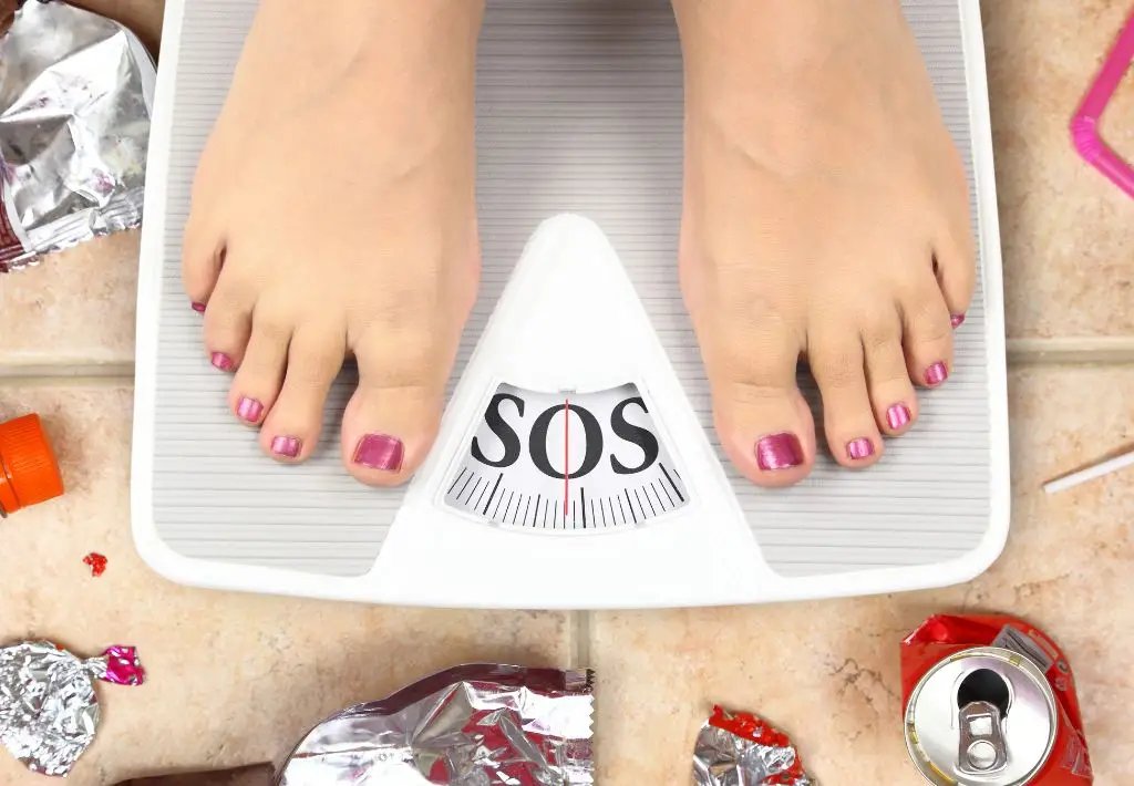 two feet with pink painted toenails stand on a white scale that says "SOS." The scale is surrounded by junkfood wrappers, suggesting that this person is in dire need of targeted weight loss programs and lifestyle changes.
