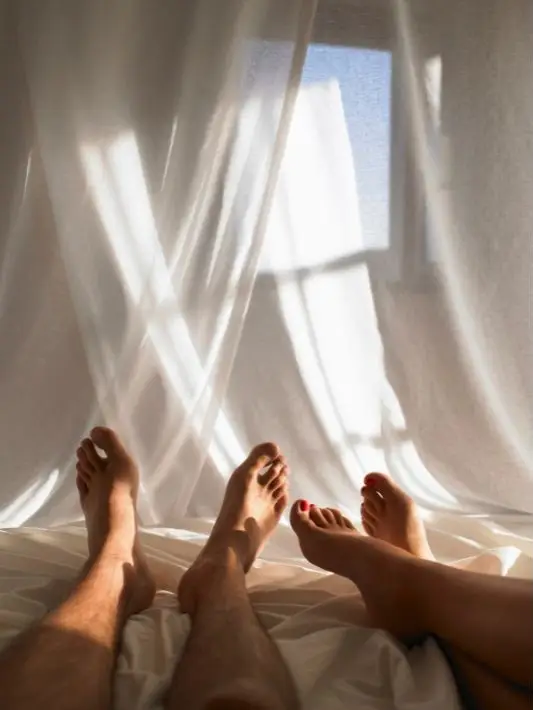 a pair of men and women's feet extend out across an umade bed while tropical white linen blows in the background.