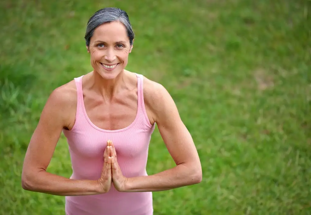 a happy older lady is in a "namaste" yoga pose as she looks up to the camera. She is smiling because she knows that anti aging for women is more than just plastic surgery. It's about inner balance and peace that comes from hormonal therapies.