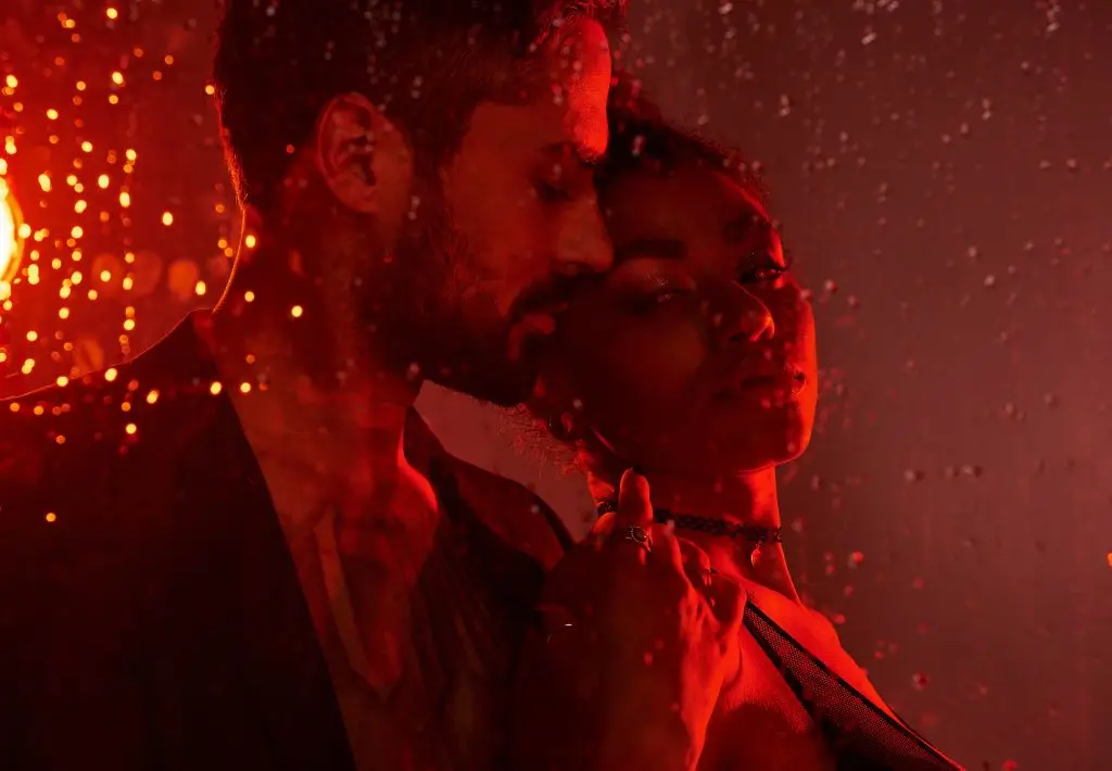 A young man and woman are backlit by a red light. She is in a little black dress and rain is on the glass that the camera is shooting through to take this picture.