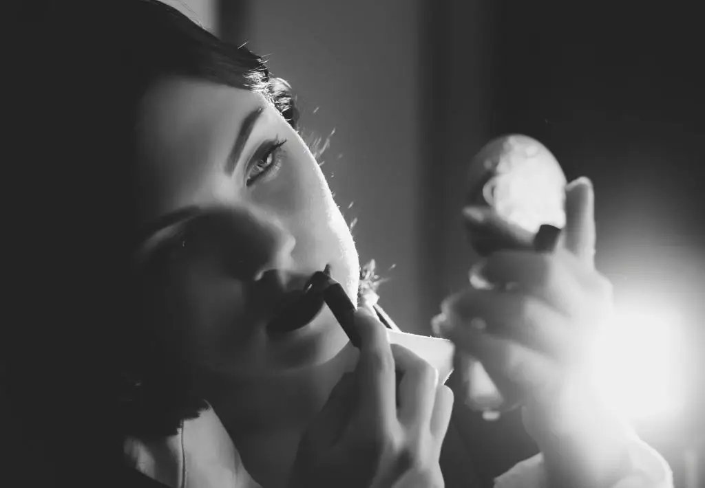 a beautifully backlit woman appears in a black and white photo as she applies red lipstic. The image calls on Elizabeth Taylor vibes - the ultimate symbol of glamorous anti aging for women.