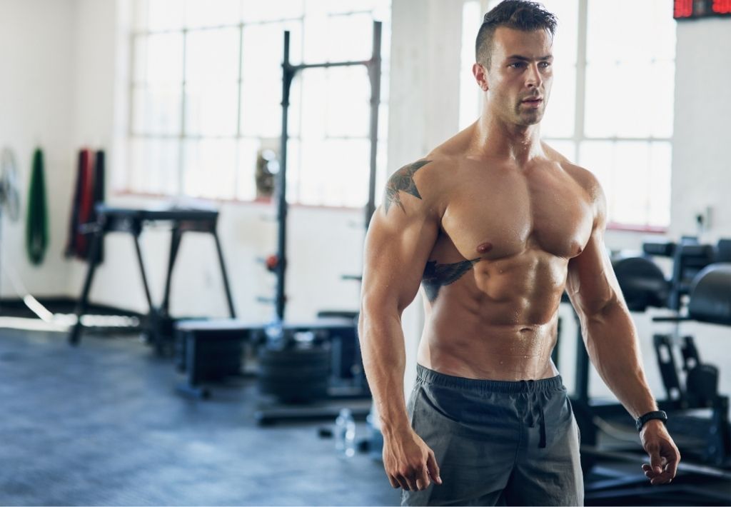 a fit man is in the gym between sets. he is without a shirt and various machines and freeweights are in the background. the average trt dose may or may not be responsible for his physique.