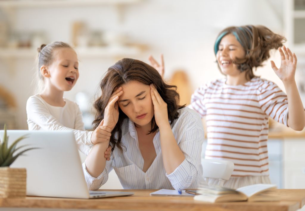 a mom rubs her temples while trying to wrok from home. her young daughters are trying to play with her but she's not feeling great due to progesterone side effects.