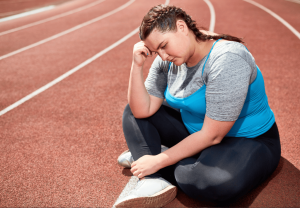 a heaviy set woman sits frustrated on a red outdoor running track. Her head is in her hands. She is wearing athletic clothes. Presumably, she cannot lose the weight and is suffering from Underactive Thyroid Weight Loss Problems.